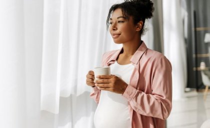 Image: Adobe. A pregnant woman holds a coffee mug and looks through sheer curtains out a window.